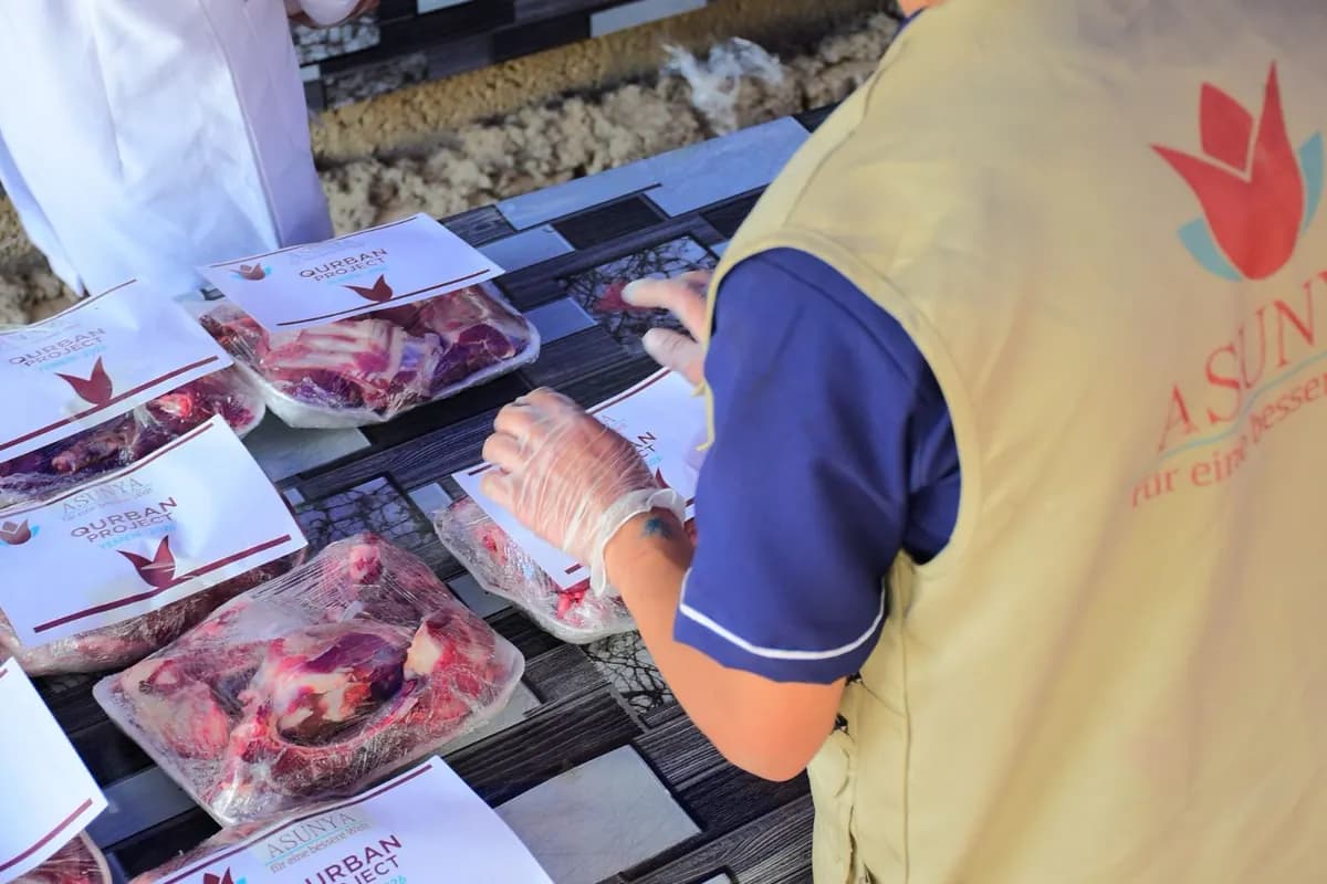 Verpackte Fleischportionen mit Etiketten des Qurban-Projekts auf einem Tisch, ein Helfer mit Handschuhen sortiert sie