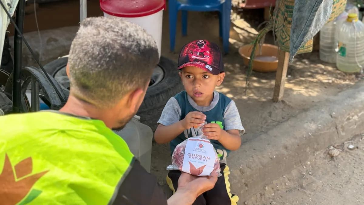 Ein Helfer in Warnweste übergibt einem kleinen Jungen mit Kappe eine Tüte mit Fleisch, auf dem Etikett steht Qurban Project
