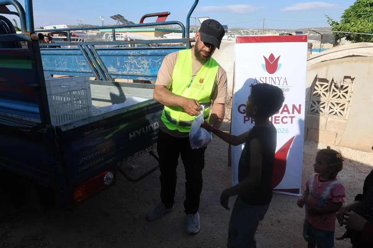 Ein Helfer in Warnweste übergibt einem Jungen eine Tüte, daneben ein Banner des Qurban-Projekts von Asunya
