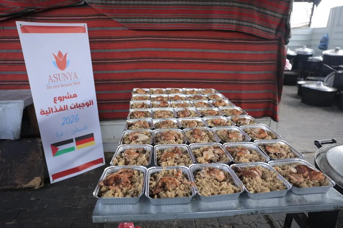 Viele Schalen mit Reis und Hähnchen stehen auf einem Tisch, daneben ein Banner von Asunya zum Mahlzeitenprojekt 2026 in Gaza.