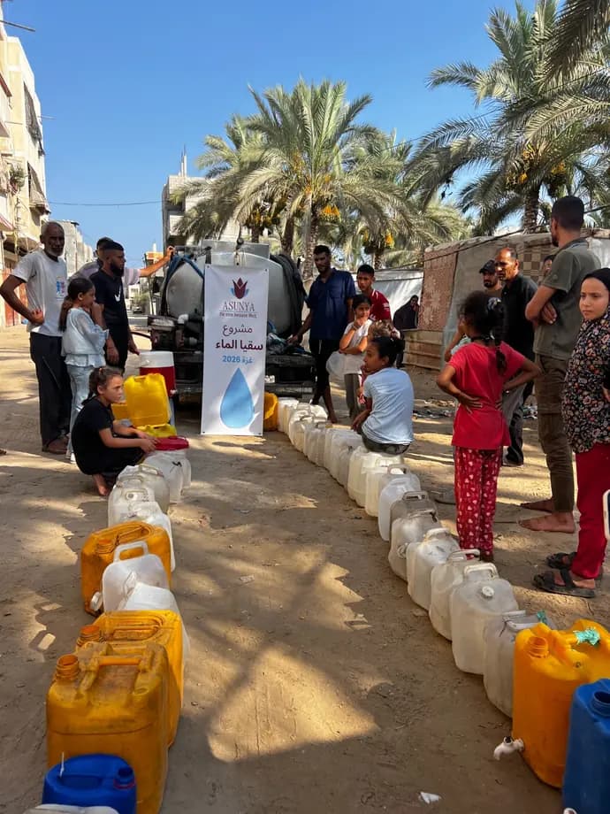 Familien stehen mit einer langen Reihe von Wasserkanistern vor einem Tankwagen mit einem Banner von Asunya zum Wasserprojekt in Gaza.
